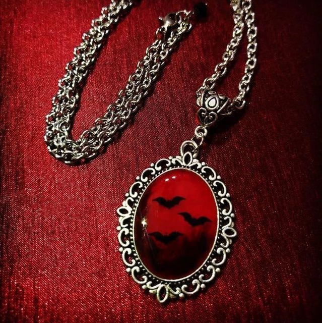 A silver-toned chain necklace with an ornate oval pendant featuring a glossy red background and black bat silhouettes, displayed on a textured red fabric surface.