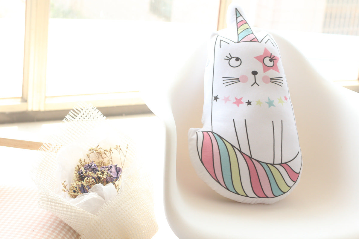 A white cat-shaped plush pillow with a colorful unicorn horn and rainbow-striped tail, styled on a white chair next to a small wrapped bouquet in a bright room.