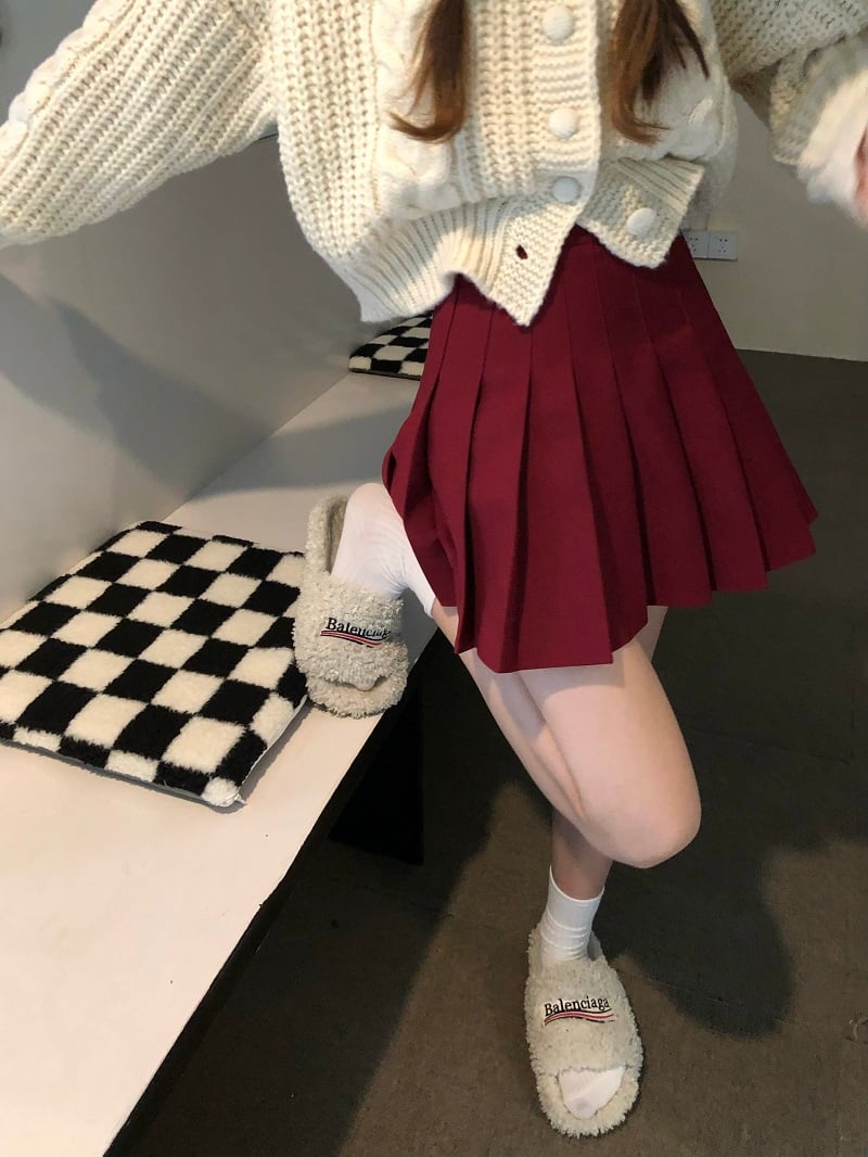 Person wearing a white knit cardigan, red pleated skirt, and white shoes in a room.