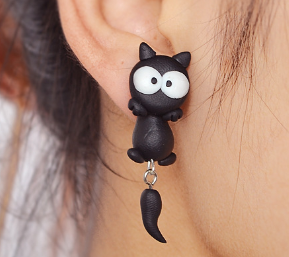 A close-up of a person's ear wearing a black cat-shaped stud earring with large white eyes and a dangling tail charm.