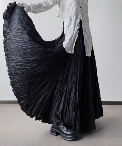A person wearing a long black pleated maxi skirt and a white top, holding out the skirt to show its volume and texture in a studio setting.