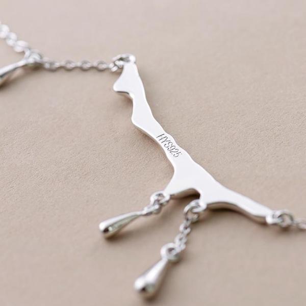 A close-up of a silver necklace featuring a giraffe silhouette pendant with two dangling teardrop charms, shown on a textured beige background.
