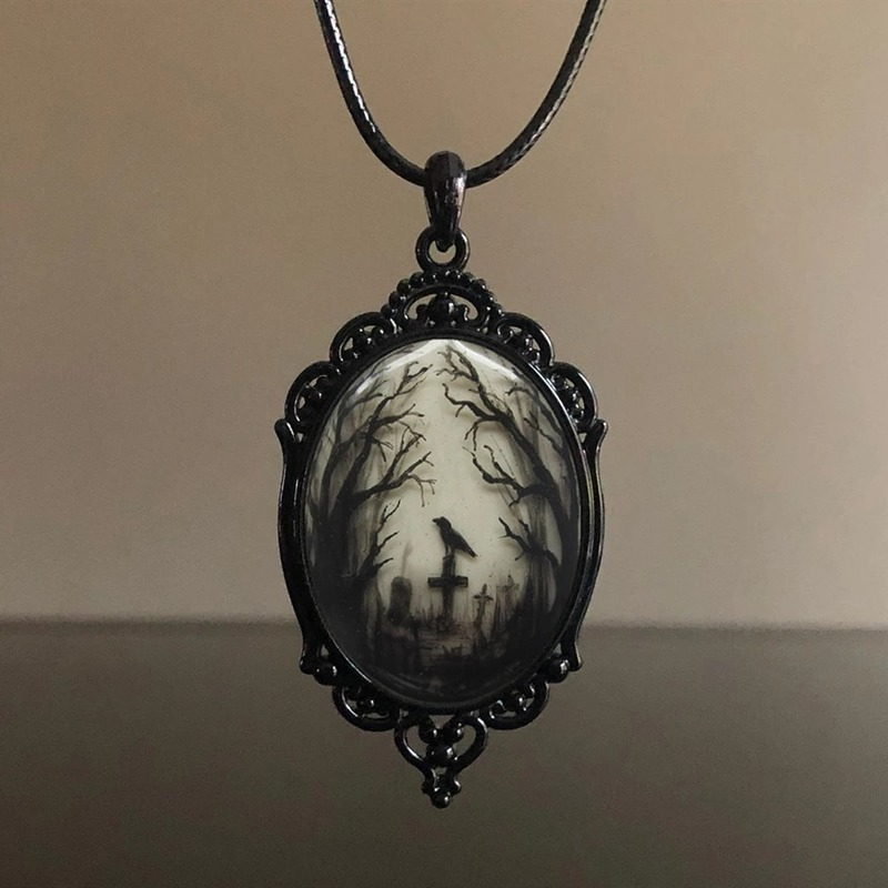 A gothic-style oval pendant necklace featuring a black metal filigree frame and a cabochon insert depicting a spooky graveyard scene with bare trees, a cross, and a raven under a pale sky, suspended from a black cord.