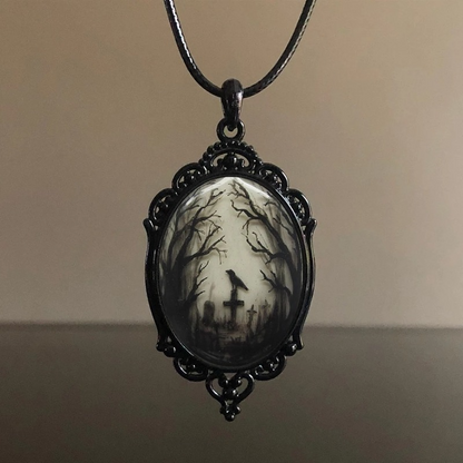 A gothic-style oval pendant necklace featuring a black metal filigree frame and a cabochon insert depicting a spooky graveyard scene with bare trees, a cross, and a raven under a pale sky, suspended from a black cord.