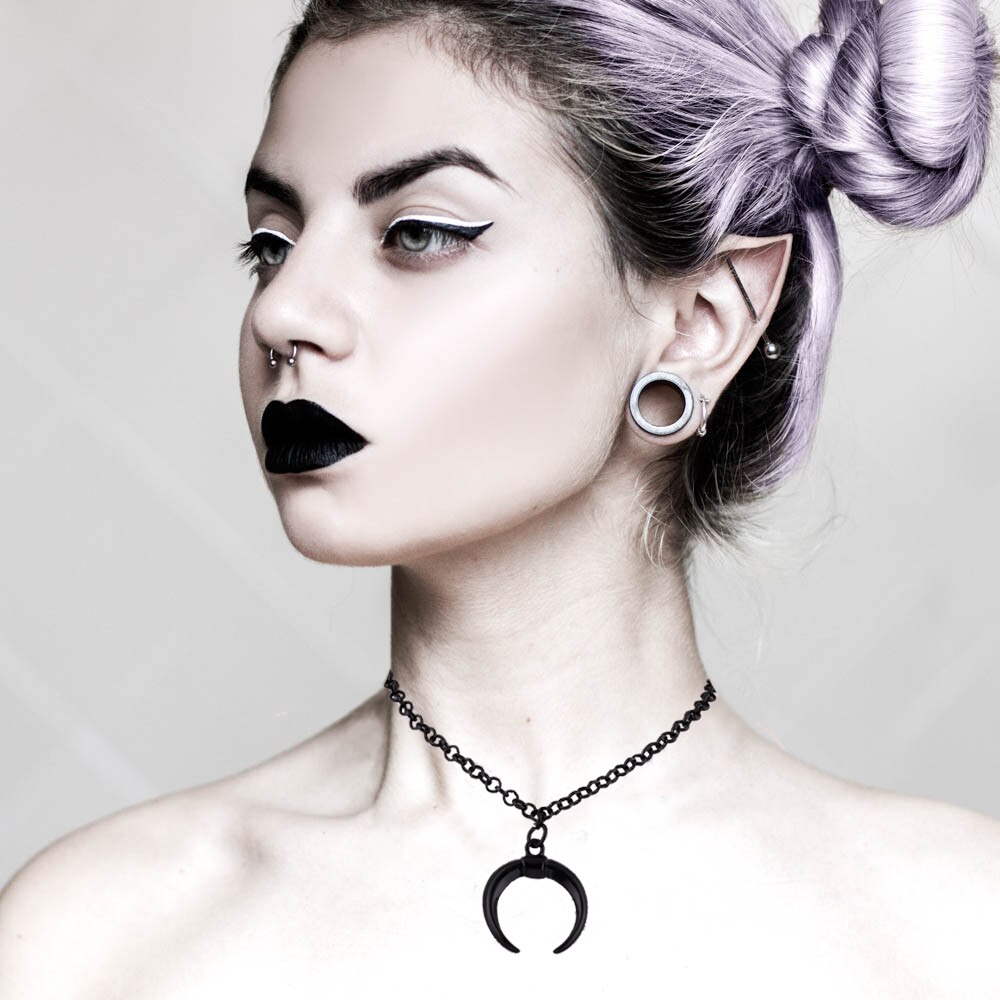 Person with black lipstick and a black crescent moon necklace against a plain background
