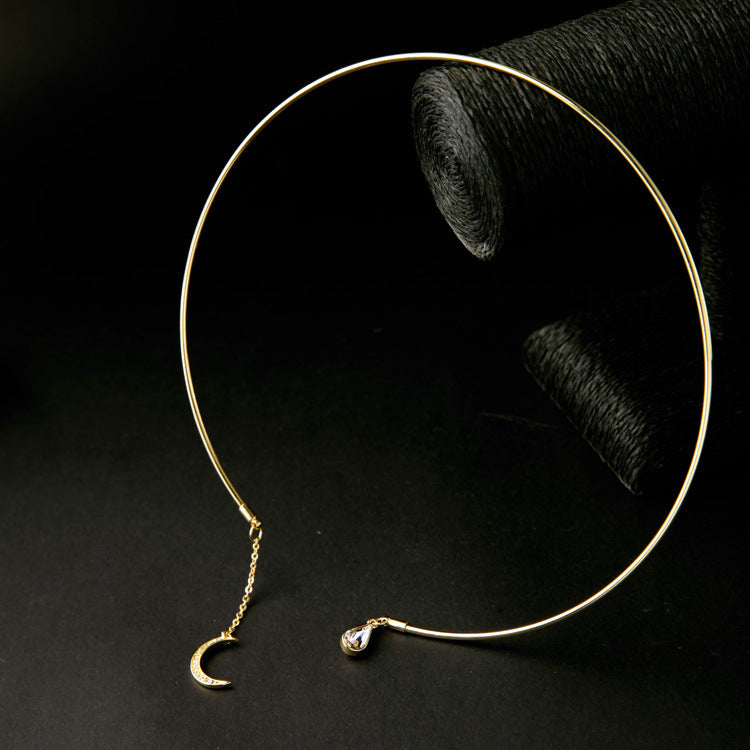 A gold-toned open hoop earring with a small crescent moon charm and a teardrop crystal accent, displayed on a black surface with a dark textured prop in the background.