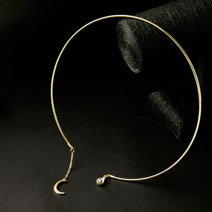 A gold-toned open hoop earring with a small crescent moon charm and a teardrop crystal accent, displayed on a black surface with a dark textured prop in the background.