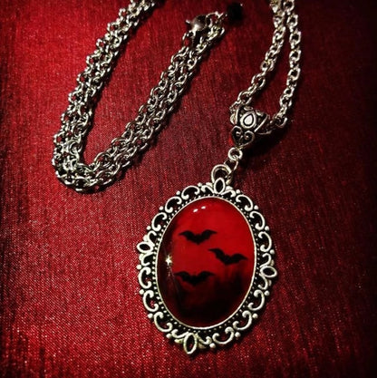 A silver-toned chain necklace with an ornate oval pendant featuring a glossy red background and black bat silhouettes, displayed on a textured red fabric surface.
