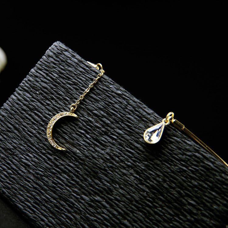 Gold-toned jewelry set featuring a crescent moon pendant with small clear stones and a teardrop-shaped clear stone charm, displayed on a textured black surface against a black background.
