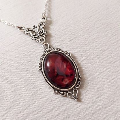 An oval pendant necklace featuring a deep red glass marbled stone set in an ornate silver-tone metal frame, hanging from a matching silver chain on a textured off-white fabric background.