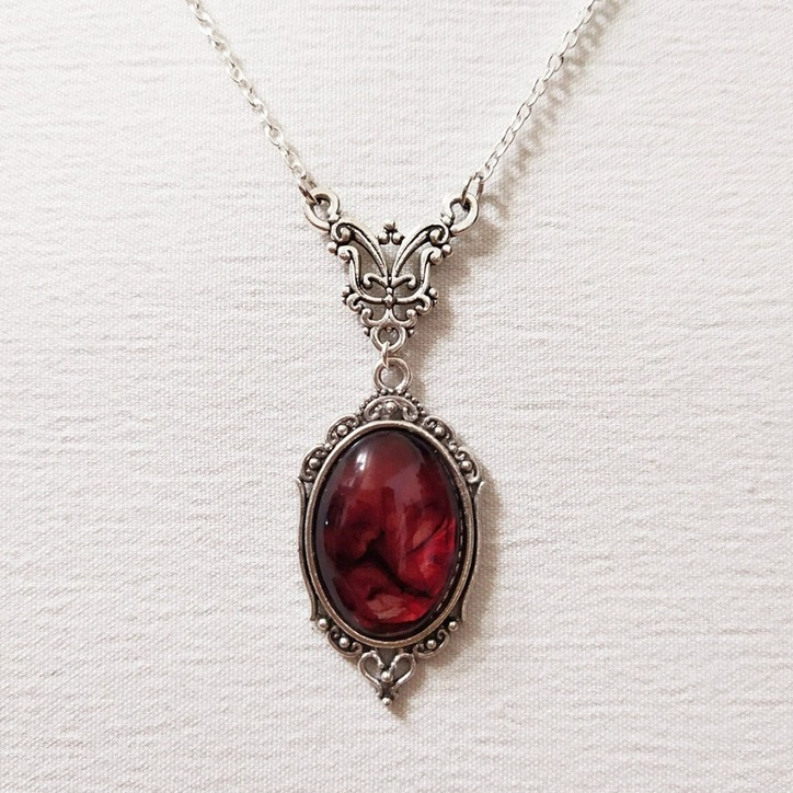 A silver-toned gothic-style pendant necklace featuring an oval, dark red marbled gemstone in an ornate filigree frame, displayed on a textured white background.