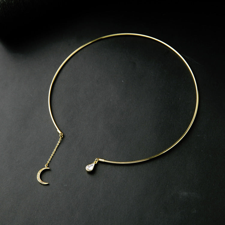 A delicate gold-toned open circle choker necklace with a small crescent moon charm and a teardrop-shaped clear stone accent, displayed on a dark textured background.