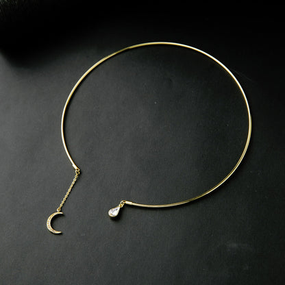 A delicate gold-toned open circle choker necklace with a small crescent moon charm and a teardrop-shaped clear stone accent, displayed on a dark textured background.