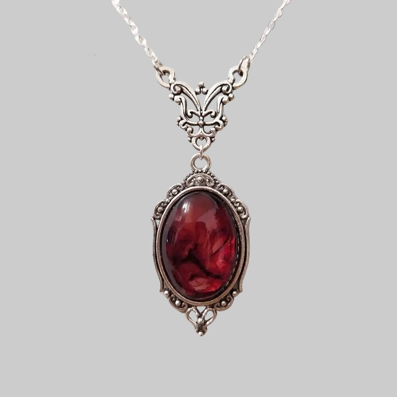 An ornate silver pendant necklace featuring a large oval red gemstone with a marbled or fluid art pattern, displayed on a textured off-white fabric background. The brand logo 'MONOYES' is visible in the top left corner.