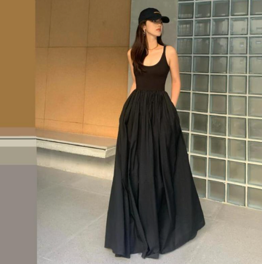 Woman wearing a black sleeveless top and long skirt with a cap, standing against a glass block wall.