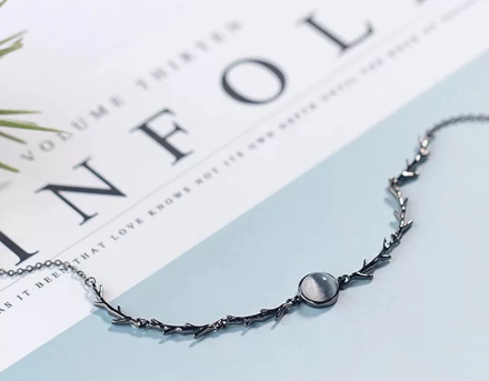A delicate silver-toned chain bracelet featuring a central round cabochon stone set within a thorny, branch-like design, displayed on a light blue surface next to a book page.
