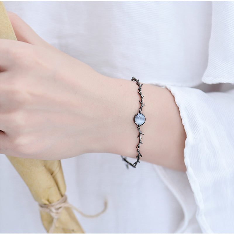 A delicate black twig-style bracelet with a round mother-of-pearl bead, worn on a person's wrist against a white shirt background.