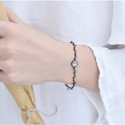 A delicate black twig-style bracelet with a round mother-of-pearl bead, worn on a person's wrist against a white shirt background.
