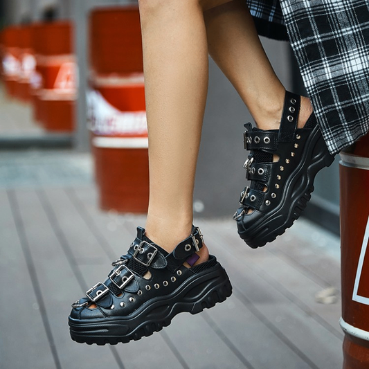 Black platform sandals with multiple silver buckles, metal eyelets, and studs, shown on a person's feet in an outdoor urban setting.