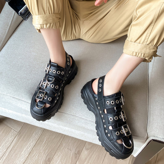 A person wearing black platform mule sandals with multiple silver-tone buckles and eyelets, seated on a light gray sofa. The person is also wearing beige rolled-up pants.