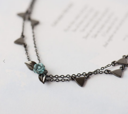 A close-up of a dark metal chain necklace featuring small triangular charms and a central turquoise-blue rose detail, resting on a white surface with blurred text in the background.
