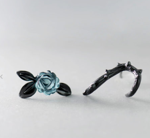 A pair of gothic-style black rings on a light gray background. One ring features a detailed light blue rose with black leaves, and the other is a thorny branch design.