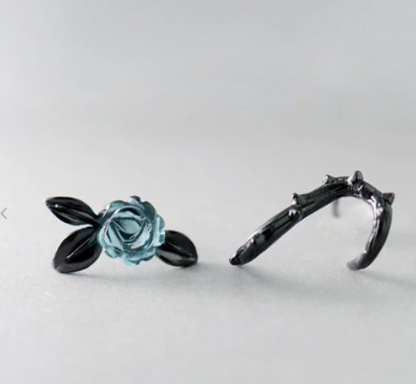 A pair of gothic-style black rings on a light gray background. One ring features a detailed light blue rose with black leaves, and the other is a thorny branch design.
