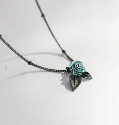 A delicate necklace featuring a small, light blue rose pendant with dark metallic leaves and a dark chain, photographed on a plain white background.