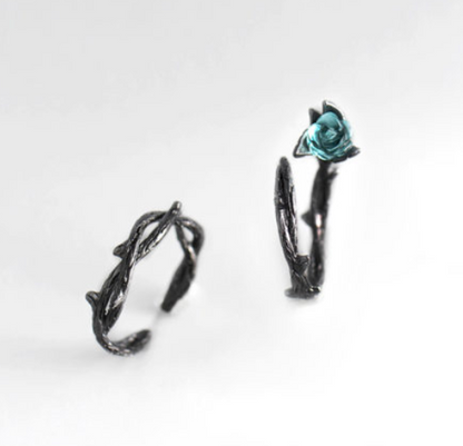 A pair of gothic-style black metal rings shaped like twisted thorny branches, with one ring featuring a small faceted teal blue rose at the top, set against a clean white background.