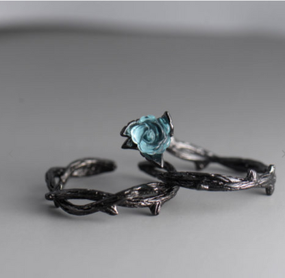 A gothic-style ring featuring a twisted barbed wire band in a dark oxidized finish, adorned with a small, vibrant light blue rose gemstone at the center, set against a neutral gray background.