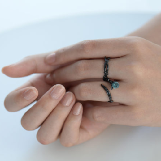A close-up of a person's hands wearing two thin black rings, one with a small round blue stone, against a soft light gray background.