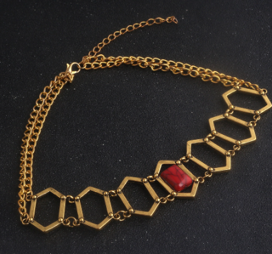 A gold-toned geometric chain necklace featuring a series of hexagonal links with a central red faceted stone, displayed on a dark textured background.