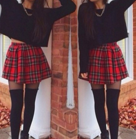 A person wearing a black cropped long-sleeve top, a red plaid pleated mini skirt, black thigh-high socks, and a silver chain necklace, standing outdoors in front of a brick column.