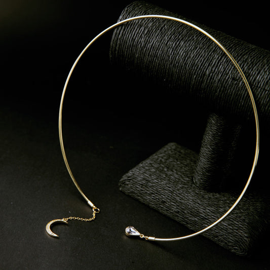 A gold-toned open circle choker necklace with a crescent moon charm and a teardrop crystal accent, displayed on a dark textured stand against a black background.