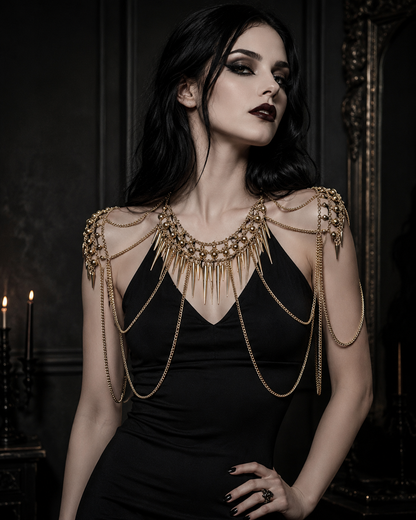 A woman with dark hair and dramatic eye makeup wears a black dress and a large ornate gold chain necklace with shoulder-mounted chains, posed in a dimly lit interior with candles and a mirror in the background.