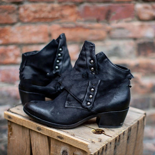 Black leather ankle boots with decorative studs on a wooden block against a brick wall.