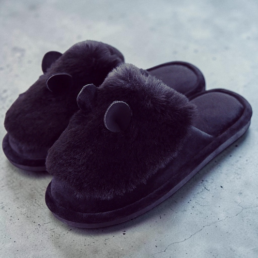 A pair of dark brown plush slippers with fuzzy bear ears on the top, photographed on a textured gray concrete surface.