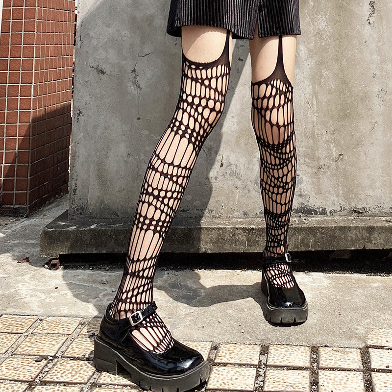 Black fishnet knee-high socks with a spiderweb pattern worn with black Mary Jane platform shoes, shown on a model's legs against a concrete wall.