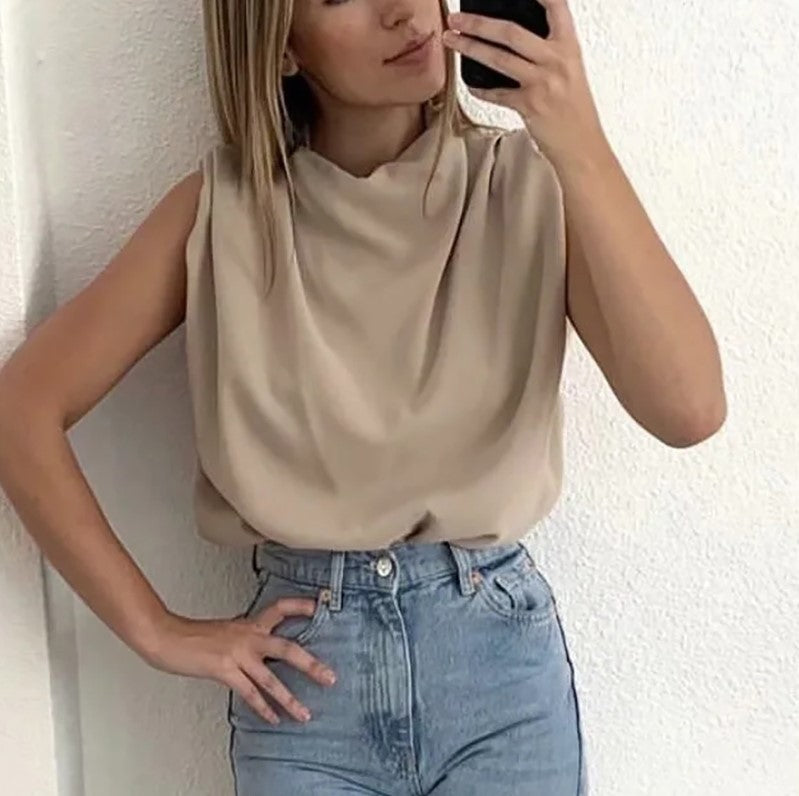 Woman wearing a beige sleeveless top and blue jeans against a white wall.