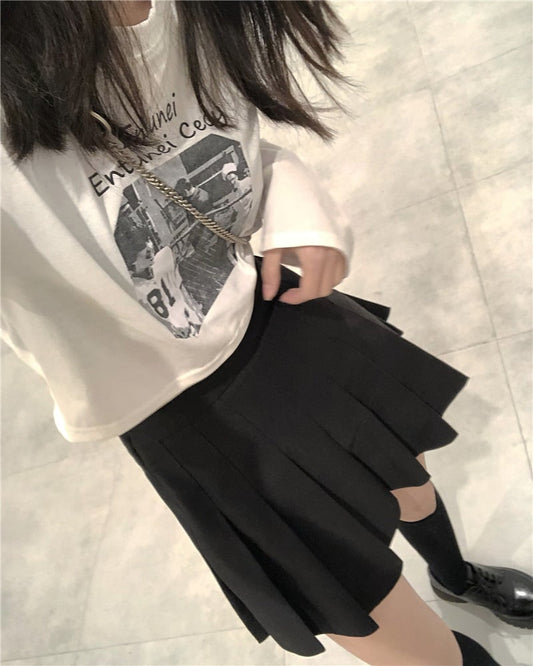Person wearing a white t-shirt with graphic design and a black pleated skirt on a light gray floor.
