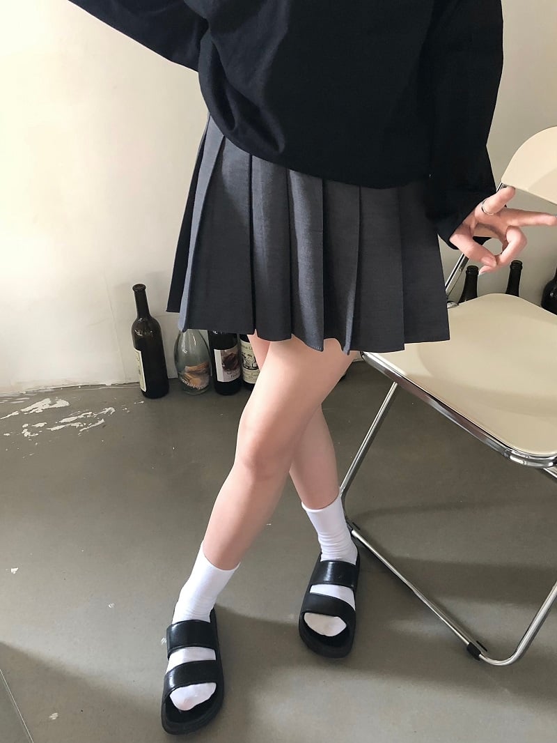 Person wearing a black sweater, pleated skirt, and black sandals in a minimal indoor setting.
