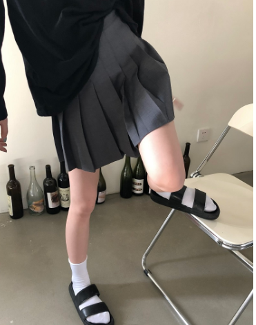 A person wearing a dark gray pleated mini skirt, a black long-sleeve top, white socks, and black-and-white slide sandals, posing with one leg raised on a white folding chair in an indoor setting.