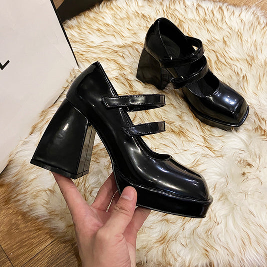Black high-heeled shoes with a strap held by a hand on a fluffy white surface.