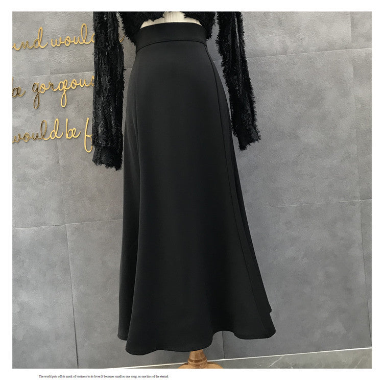 A black high-waisted A-line midi skirt displayed on a mannequin torso, paired with a black fuzzy long-sleeve top, set against a gray tiled wall with gold script lettering.