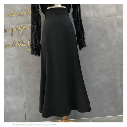 A black high-waisted A-line midi skirt displayed on a mannequin torso, paired with a black fuzzy long-sleeve top, set against a gray tiled wall with gold script lettering.