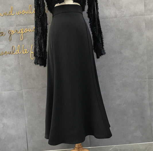 A black high-waisted A-line midi skirt displayed on a mannequin torso, paired with a black fuzzy long-sleeve top, set against a gray tiled wall with gold script lettering.