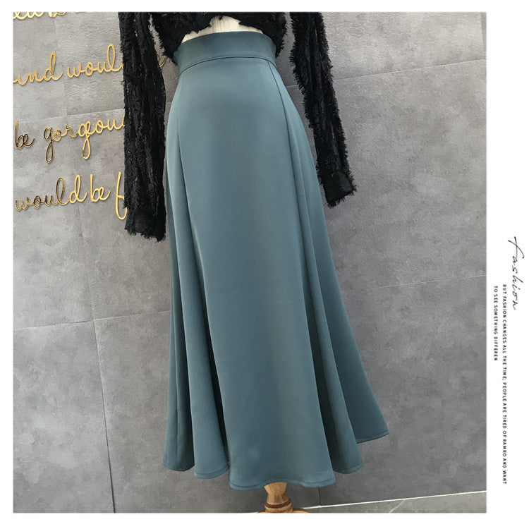 A high-waisted, A-line midi skirt in a muted slate blue color, displayed on a mannequin torso with a black long-sleeve top. The background is a gray textured wall with gold decorative lettering.