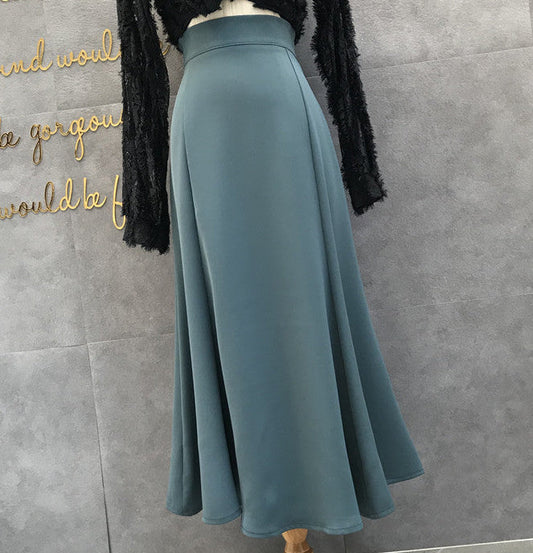A high-waisted, A-line midi skirt in a muted slate blue color, displayed on a mannequin torso with a black long-sleeve top. The background is a gray textured wall with gold decorative lettering.