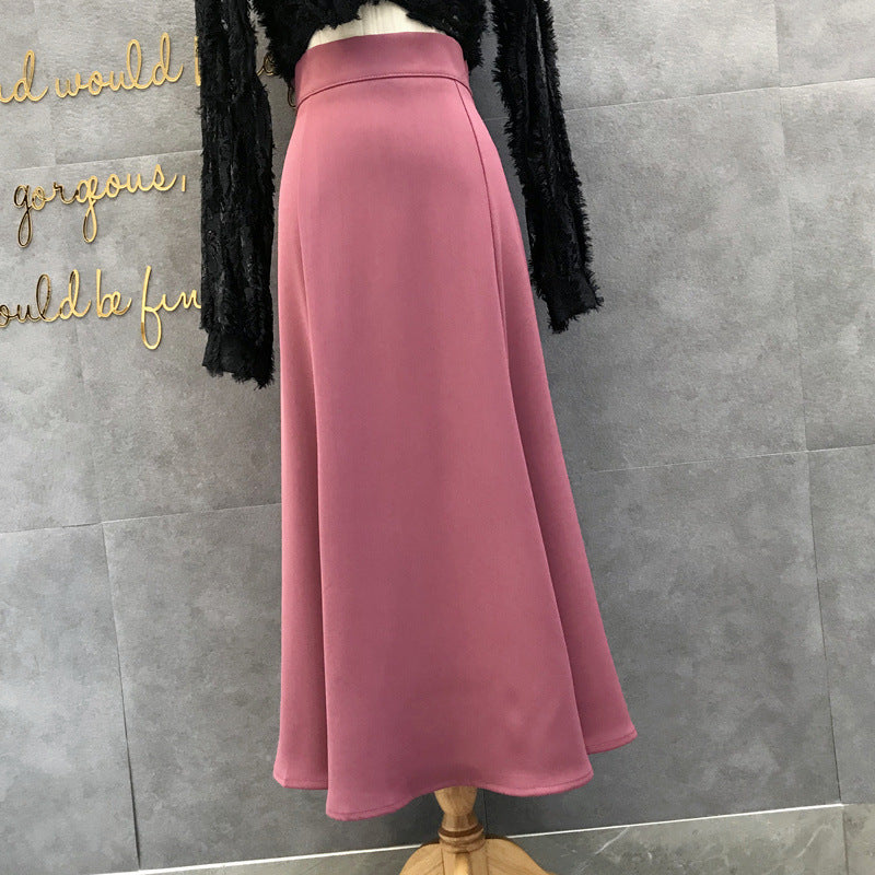 A dusty rose pink high-waisted A-line midi skirt displayed on a mannequin torso, paired with a black fringed cardigan, set against a gray tiled wall with gold script lettering.