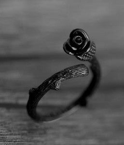 A dark metal ring featuring a textured branch-like band and a rose-shaped top with a small central stone, shown in black and white on a wooden surface.